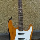 Elektrická kytara s žlutohnatým trupem a hnědým hmatníkem. Na hlavě je viditelné logo Fender. Kytara má bílý pickguard, tři snímače a kovový most s i…