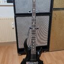 Černá elektrická basová kytara s vyřezávaným korpusem, umístěná na stojanu před dvěma komby s nápisem CANON. Na hlavě nástroje je logo Ltd. Hrabednic…