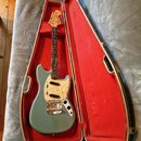 Modrá elektrická kytara v otevřeném tvrdém kufru s červeným vnitřkem. Na hlavě hmatníku je viditelné logo Fender. Kytara má bílý pickguard a chromova…