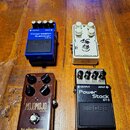 Na dřevěném povrchu jsou vedle sebe čtyři efektové pedály. Vlevo nahoře je modrý pedal s nápisy BOSS, Compression Sustainer CS-3, OUTPUT a INPUT. Vpr…