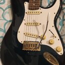 Tmavě modrá elektrická kytara typu Stratocaster s bílým pickguardem a hnedým hmatníkem. Nástroj má tři snímače, ladicí knofíky a hardware v zlatém pr…