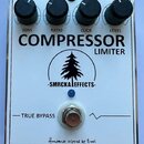 Bílý efekt pedál s nápisy COMPRESSOR a LIMITER. Na horní hraně jsou čtyři černé oválné knoflíky s popisky SENS, RATIO, CLICK a LEVEL. Uprostřed je lo…