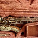 Zlatý lakovaný saxofon v hnědém polstrovaném kufru. Nástroj má bílé klapkové polštáře a v kufru je vedle něj položen mouthpiece. Na těle nástroje nej…