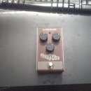 Obdélníkový efekt pedál v hnědé barvě s nápisy tc electronic a Rusty FUZZ. Na horní straně jsou tři černé ovládací knoflíky a červená kontrolka. V do…