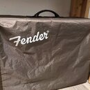 Hnědý polstrovaný obal na hudební nástroj s černou rukojetí. Na přední straně je bílý nápis Fender®. Povrch materiálu vykazuje výrazné pomačkání a vr…