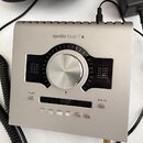 Stříbrné audio rozhraní s nápisem apollo twin | x. Uprostřed se nachází velký kovový ovládací knoflík obklopený stupnicí a dvěma LED indikátory úrovn…