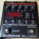 Černý efekt pedál s nápisem t.c. electronic a označením ND-1 NOVA DELAY PROGRAMMABLE DIGITAL DELAY. Horní část obsahuje pět ovládacích knoflíků s pop…