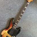 Elektrická basová kytara s dřevem hmatníku a tělem v přechodovém lakování od černé přes červenou po žlutou. Na hlavě je nápis "Fender JAZZ BASS". Čer…
