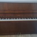 Hnědé dřevěné piano s klávesnicí. Na přední horní liště nad klávesami je zlatý nápis PETROF. Nástroj vykazuje známky běžného používání, povrch dřeva…