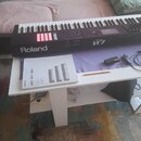 Černý digitální klavír s bílými a černými klávesami. Na přední straně je bílý nápis Roland a modelové označení FA-07. Vedle klavíru leží bílý manuál…