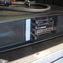 Černý rackový zesilovač s kovovým perforovaným předním panelem. Na ovládacím panelu jsou čtyři otočné ovladače a nápis FP 20000Q. Zařízení je umístěn…