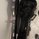 Otevřený černý tvrdý kufýr na kytaru s vnitřním polstrováním z černého plyšového materiálu. Na vnitřní straně je vidět nápis "Fender". Kufýr má červe…