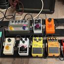 Pedálboard s osmi kytarovými efekty rozmístěnými ve dvou řadách. V horní řadě jsou pedály s nápisy „electro-harmonix“, „MXR phase 90“ a jeden zelený…