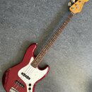 Čtyřstrunná elektrická basová kytara s červeným metallic povrchem těla a bílým pickguardem. Na hlavě hmatníku je viditelné logo Fender Japan. Hmatník…