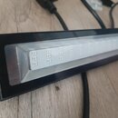 Černý obdélníkový LED panel s bílým svítícím polem, které obsahuje mřížku malých světelných bodů. Zařízení je připojeno k černému napájecímu kabelu.…