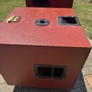 Červený kubický reproduktorový box s texturovaným povrchem. Na horní straně je kovová rukojeť, kulatý otvor a jeden obdélníkový basreflexní port. Na…