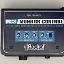 Tmavě modrý studio monitor controller s bílým nápisem MC3 MONITOR CONTROL a STUDIO MONITOR CONTROLLER. Zařízení obsahuje čtyři ovládací knoflíky s po…