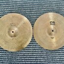 Dva mosazné činely typu hi-hat položené vedle sebe na modrém koberci. Levý činel nese nápisy PAISTE 2002 a HI-HAT BOTTOM, pravý činel nápisy PAISTE 2…