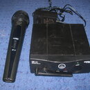 Černý bezdrátový mikrofon a odpovídající přijímač s napájecím adaptérem. Na mikrofonu je bílý nápis MINI PRO a HT 40, na hlavě logo AKG. Přijímač nes…