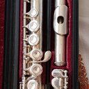 Stříbrná flétna rozložená v černém kufru s červeným polstrováním. Na horní části hlavního trupu je vidět nápis YAMAHA a pod ním menší, nečitelný text…
