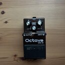 Černý efekt pedál s nápisy Octave a OC-2. Na horní části jsou tři ovládací knoflíky, červená kontrolka s nápisem CHECK a popisy DIRECT LEVEL, OCT 2 a…