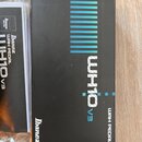 Černá papírová krabice s modrými pruhy a bílým nápisem WH10v3 WAH PEDAL. V levém dolním rohu je bílé logo Ibanez. Vedle krabice je položen papírový m…