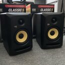 Dva černé studiové monitory s žlutými membránami basových reproduktorů. Na obou monitorech je bílé logo KRK. V pozadí jsou dvě krabice s nápisy KRK,…