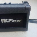 Černý malý kombozesilovač s texturovaným povrchem a plastovými rohy. Na přední mřížce je bílý nápis STARSound. K přístroji je připevněn černý tkaný p…