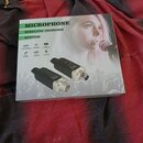 Fotografie znázorňuje bílé papírové krabici s produktem. Na krabici je nápis MICROPHONE WIRELESS CHARGING SYSTEM a obrázek ženy s mikrofonem. V dolní…