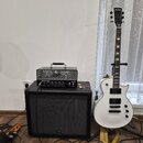 Bílá elektrická kytara s černým hmatníkem a dvěma humbuckery, na hlavě je nápis Hartke Bass. Vedle ní stojí černý kytarový kombozesilovač s logem Har…