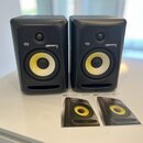 Dva černé studiové monitory s žlutými membránami basových reproduktorů. Na obou je vidět logo KRK a nápis ROKIT 6. Před reproduktory leží dva papírov…