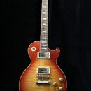 Elektrická kytara s trupem ve tvaru Les Paul v červeně-žlutém sunburst provedení s viditelnou strukturou dřeva. Na hlavě hmatníku je bílý nápis Gibso…