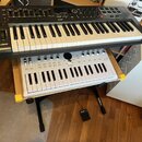 Na fotografii jsou dvě klaviatura umístěná na stojanu. Horní černá klaviatura nese nápisy novation a IMPULSE. Spodní bílá klaviatura má nápis ARTURIA…