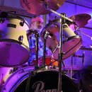 Bílá bicí souprava značky Pearl s černým basovým bubnem, na kterém je bílý nápis Pearl. Souprava obsahuje dva tom-tomy a činely značky Zildjian. Za n…