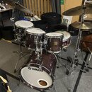 Akustická bicí souprava v tmavě hnědé barvě s viditnou strukturou dřeva. Na předním resonantním obruči basového bubnu je černé logo a nápis YAMAHA. S…