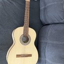 Klasická kytara s přírodním dřevěným povrchem korpusu a hnědým hmatníkem. Na desce u ozvukové otvoru je nápis „la Guitarra“. Kolem ozvukového otvoru…