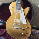 Zlatá elektrická kytara s tvarem single-cut v černém kufru s purpurovým vnitřkem. Nástroj má dva humbuckery s krémovými kryty, bílý plechtr zaštitu a…