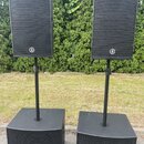 Dva sety černých reproduktorů, každý tvořený horním menším reproduktorem na stojanu a spodním větším subwooferem. Na předních mřížkách všech čtyř rep…