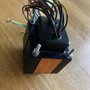 Černý kovový box s chladicími žebry a měděným prvkem na přední straně. Z horní části vedou svazky barevných elektrických vodičů. Zařízení je upevněno…