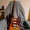 Elektrická kytara s trupem v barevnosti sunburst a hnědým hmatníkem. Kytara má červeně mramorovaný pickguard a na hlavě je viditelné logo Fender. Pov…