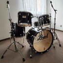 Černá bicí souprava značky Pearl s chromovaným kováním. Na přední straně basového bubnu je velký nápis Pearl, na tom-tamech jsou vidět menší loga Pea…