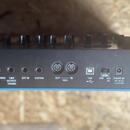 Detail zadní strany černého elektronického hudebního nástroje s řadou konektorů. Jsou zde vidět vstupy a výstupy s popisky HEADPHONES, LINE OUTPUT (M…