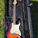 Elektrická kytara s korpusem v barevnosti sunburst a bílým pickguardem leží v černém polstváném kufru. Nástroj má hmatník z tmavého dřeva a světle hn…