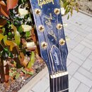 Fotografie zachycuje hlavu a část hmatníku elektrické kytary v černé barvě. Na hlavě je zlatým písmem nápis Gibson a dále označení Les Paul MODEL Stu…