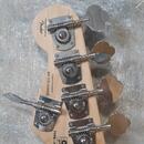 Fotografie zachycuje hlavu basové kytary z přírodního dřeva se čtyřmi kovovými ladičkami. Na dřevě jsou nápisy „Designed and licensed by Fender“, „Cr…