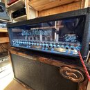 Kytarový zesilovač s černým krytem a proskleným předním panelem, pod kterým jsou vidět elektronické lampy. Na skle je bílý nápis Hughes & Kettner a v…