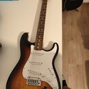 Elektrická kytara typu Stratocaster s barevným přechodem korpusu od černé přes hnědou k žluté. Má bílý pickguard, tři snímače a hnědý hmatník. Na hla…
