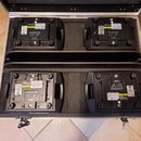 V otevřeném hliníkovém kufru s pěnovou výplní jsou umístěny čtyři černé kvadratické jednotky elektronického vybavení. Na spodních stranách zařízení j…
