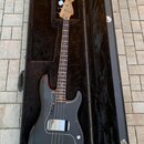 Černá elektrická basová kytara v černém tvrdém kufru. Na hlavě hmatníku je nápis Fender. Nástroj má tmavý hmatník, bílý pickguard a chromované hardwa…