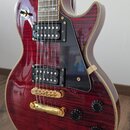Elektrická kytara s červeným plamenovitým povrchem a zlatými komponenty. Nástroj má dva humbuckery s černými kryty, černý pickguard a čtyři černé ovl…